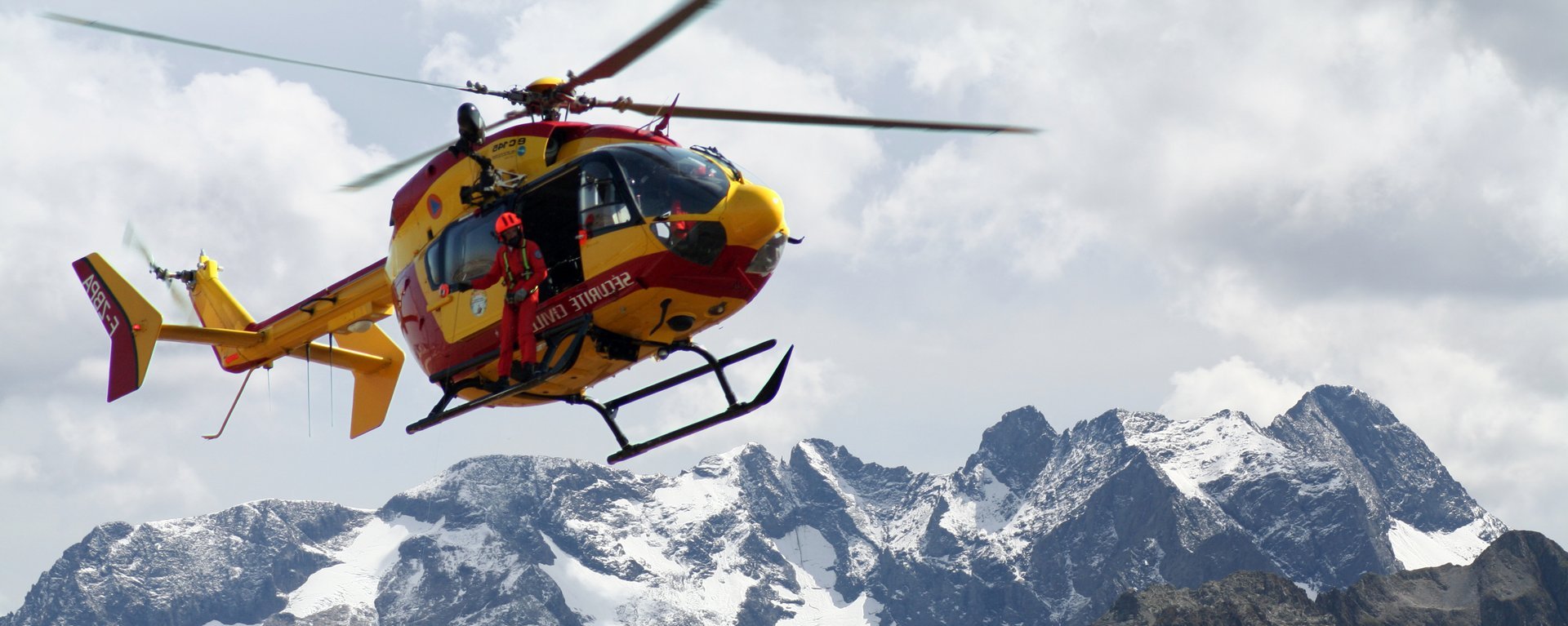 Ein Bergrettungshubschrauber mit einem Rettungshelfer fliegt über Gebirge. Der Hubschrauber im Einsatz ist im oberen linken Bildviertel platziert und befindet sich vor einem schönen Bergpanorama mit schneebedeckten Gipfeln bei einem hellen, mit Wolken bedeckten Himmel.