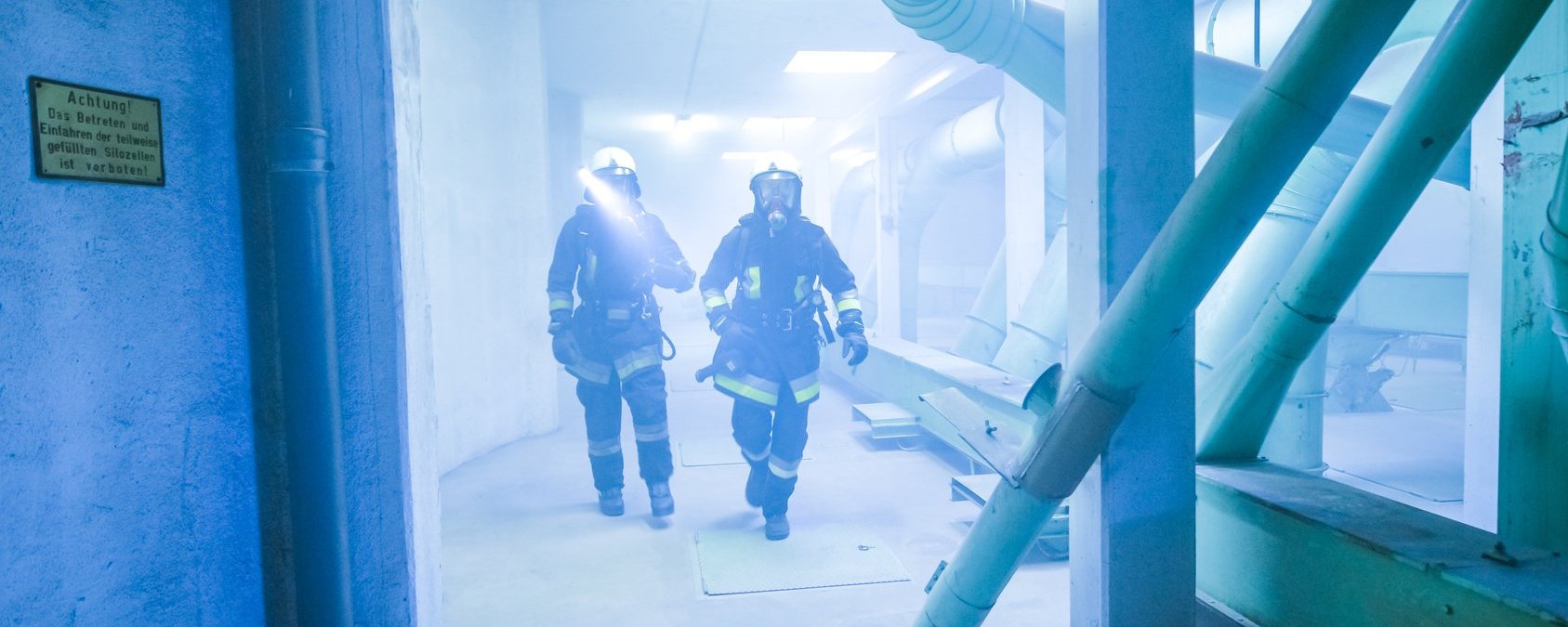 Zwei Feuerwehrmänner befinden sich in einem verrauchten Industriegebäude mit Rohren und Betonsäulen. Die beiden Personen sind in voller Einsatzkleidung und laufen mit Licht und Atemmasken auf die Kamera zu.