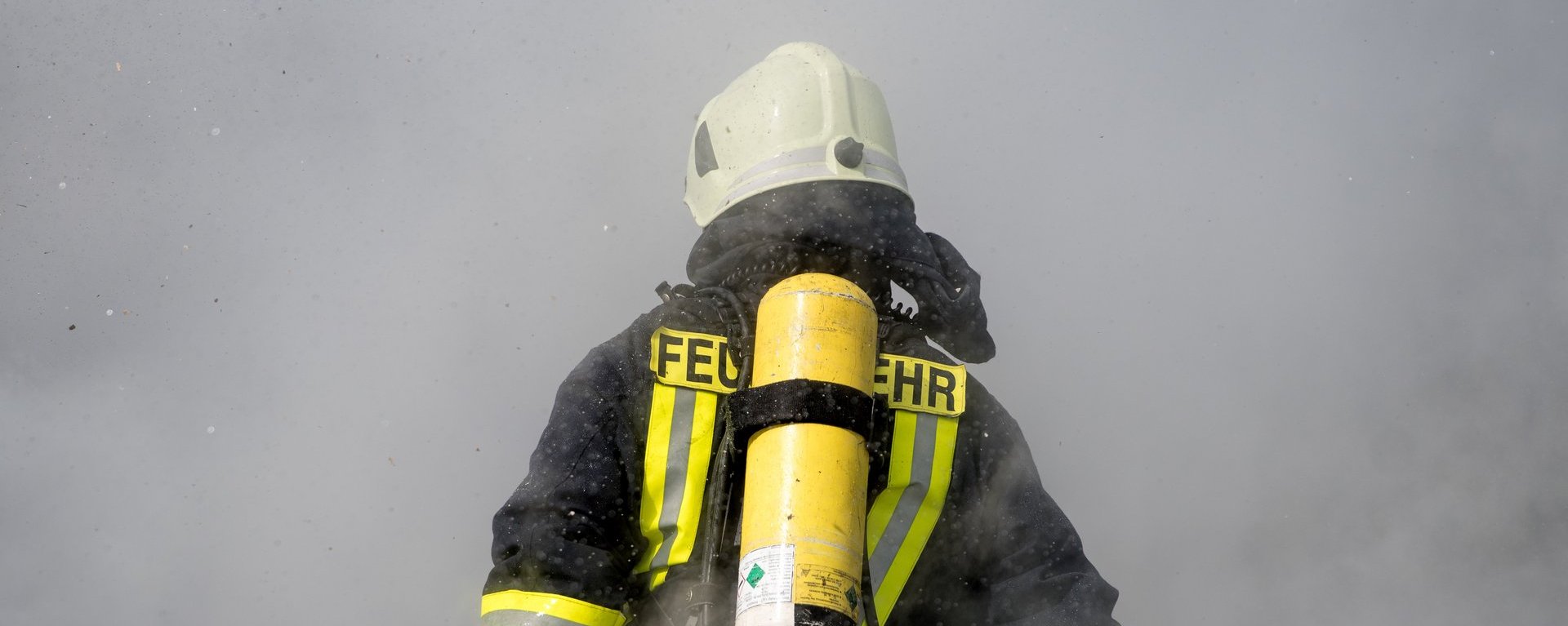 Rückansicht eines Feuerwehrmanns in Ausrüstung der in eine Rauchschwade hineinläuft. Mit Helm auf dem Kopf und Sauerstoffflasche auf dem Rücken wird sein Unterkörper bereits komplett vom Rauch umhüllt.