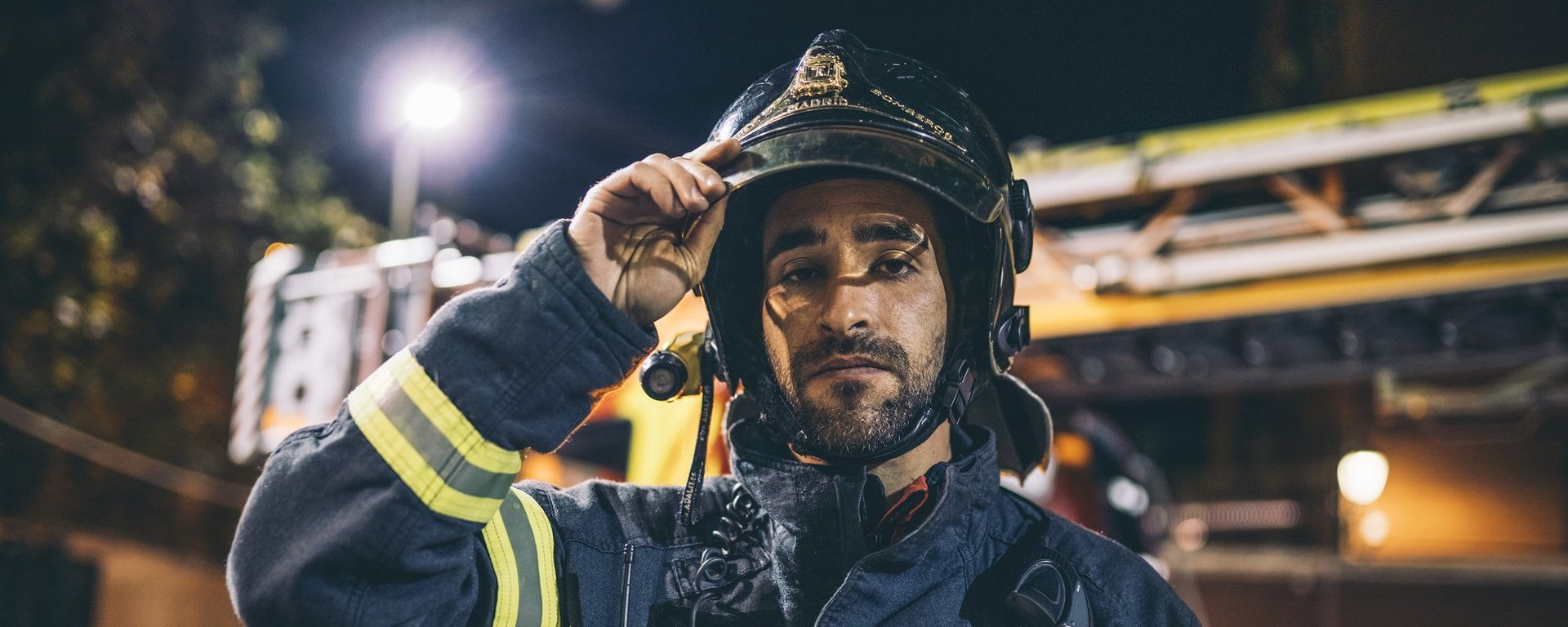 Frontalaufnahme eines Feuerwehrmanns in Einsatzklamotten, der in die Kamera blickt und sich an das Visier seines Helmes fasst. Im Hintergrund ist ein Feuerwehrlagen mit Drehleiter auf dem Dach zu sehen.