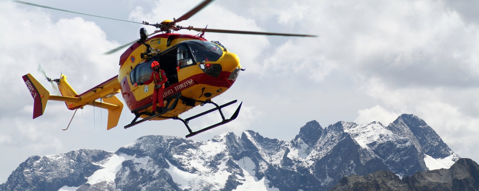 Ein Bergrettungshubschrauber mit einem Rettungshelfer fliegt über Gebirge. Der Hubschrauber im Einsatz ist im oberen linken Bildviertel platziert und befindet sich vor einem schönen Bergpanorama mit schneebedeckten Gipfeln bei einem hellen, mit Wolken bedeckten Himmel.