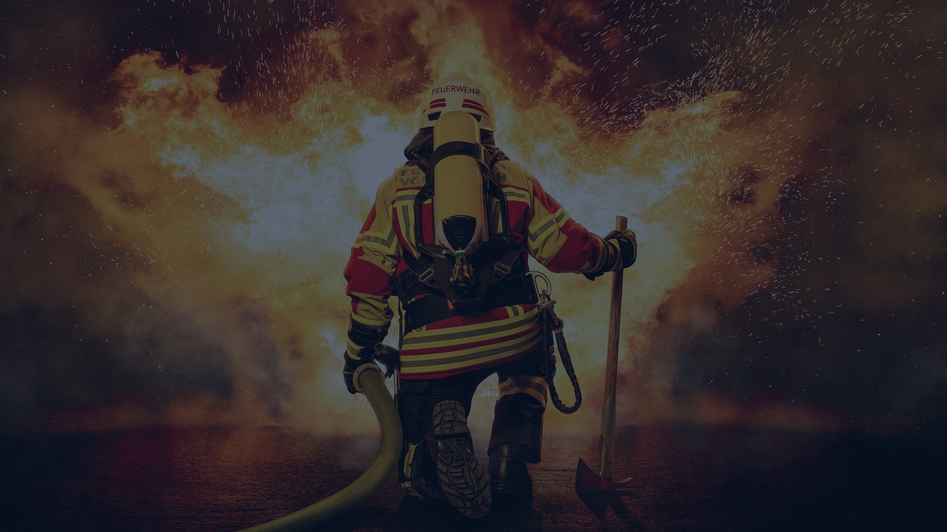 Ein Feuerwehrmann mit Schutzkleidung, Sauerstoffflasche, Beil und Wasserschlauch ist von hinten zu sehen und einem großen, lodernden Feuer zugewandt. Das Bild wurde mit einer vollflächigen dunkelblauen Überlagerung abgedunkelt.