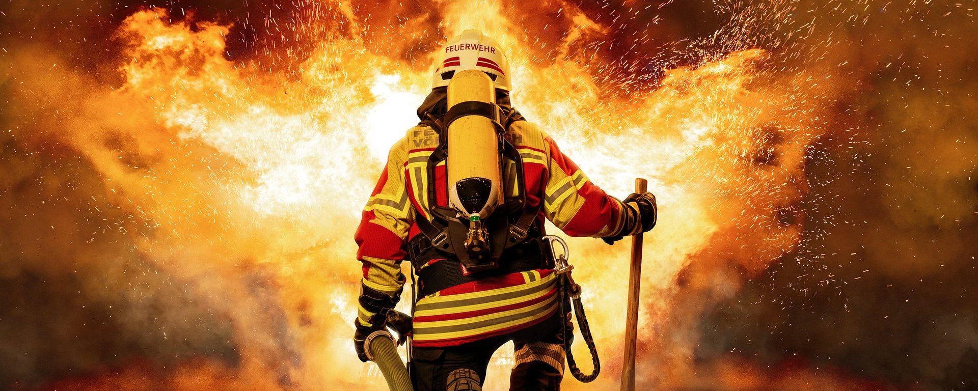 Ein Feuerwehrmann mit Schutzkleidung, Sauerstoffflasche, Beil und Wasserschlauch ist von hinten zu sehen und einem großen, lodernden Feuer zugewandt.